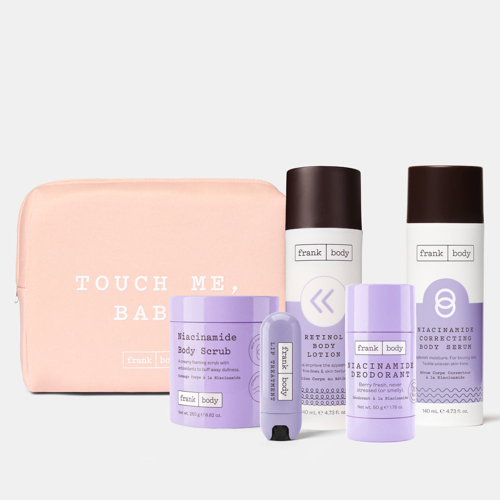 A set of Frank Body personal care products arranged in front of a pale pink zip pouch on a light gray background. The set includes a pink pouch with white text reading 'TOUCH ME, BABY', a lavender body scrub jar, a white body lotion bottle with a dark cap, a lavender deodorant stick, and a white body wash bottle with a dark cap.