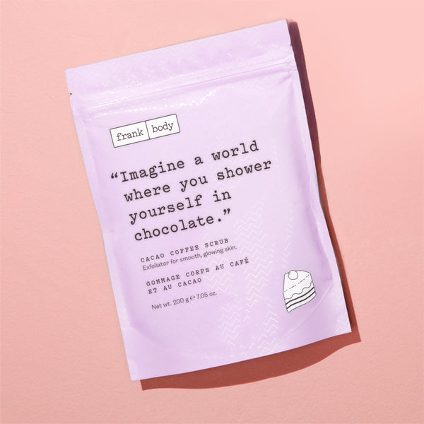 A lavender stand-up pouch of Frank Body Cacao Coffee Scrub, featuring a printed quote and a small cake illustration, set against a solid pink background.