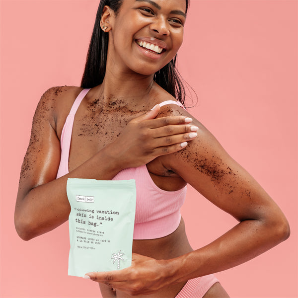 A smiling woman with dark charcoal powder on her skin holds a mint green pouch of Coconut Coffee Scrub against a solid pink background.
