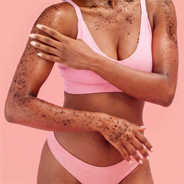 A woman wearing a light pink ribbed bikini top and bottom, with dark charcoal scrub applied all over her skin, posed against a solid pink background.