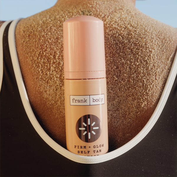 A bottle of frank body Firm + Glow Self Tan Mousse held against a bare back with tan mousse applied to the shoulders.