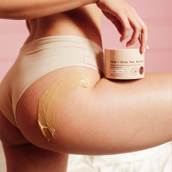 A woman in nude underwear holding a tub of frank body Firm + Glow Tan Butter beside her hip, the butter smoothed onto her skin.