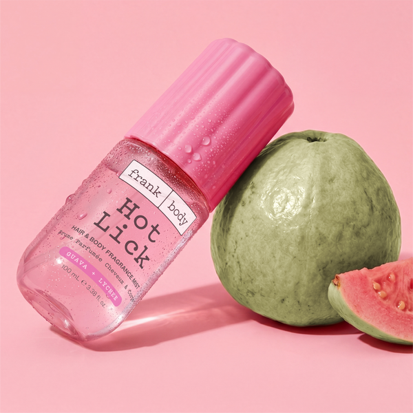 A bottle of frank body Hot Lick Hair & Body Mist leaning against a whole and halved guava on a pink background.