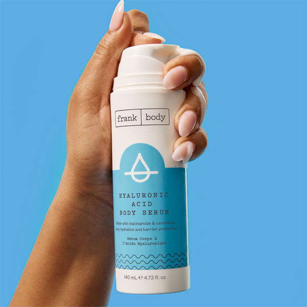 A hand holding a bottle of frank body Hyaluronic Acid Body Serum against a blue background.