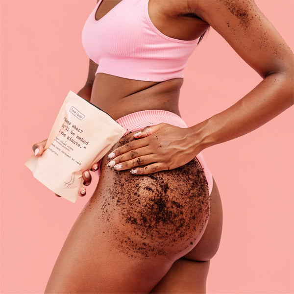 A woman in a pink ribbed sports bra and matching shorts holds a beige paper pouch labeled 'Original Coffee Scrub' while applying a dark brown scrub to her upper thigh. The background is a solid pastel pink.