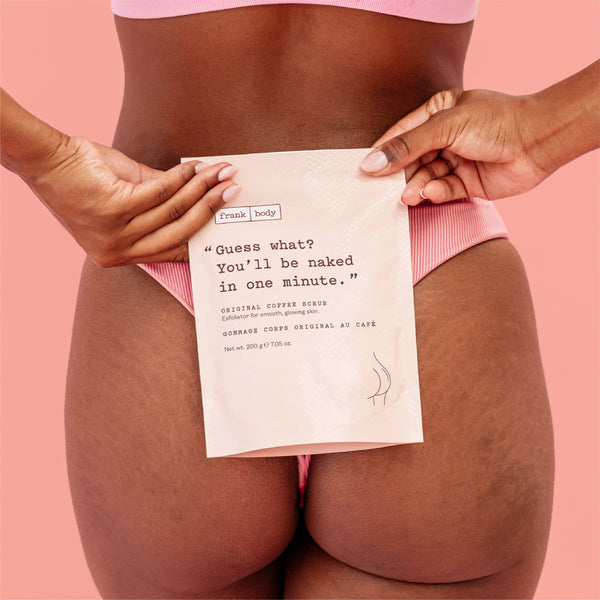 A person wearing pink underwear holds a white pouch of Frank body Original Coffee Scrub against their lower torso, with the text on the pouch reading, "Guess what? You'll be naked in one minute." against a solid pink background.