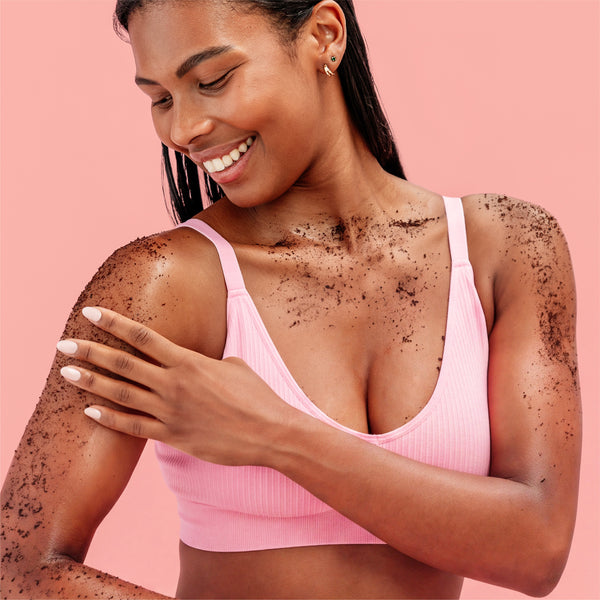 A woman with dark brown body scrub applied to her skin, wearing a light pink ribbed sports bra, posing against a solid pink background.