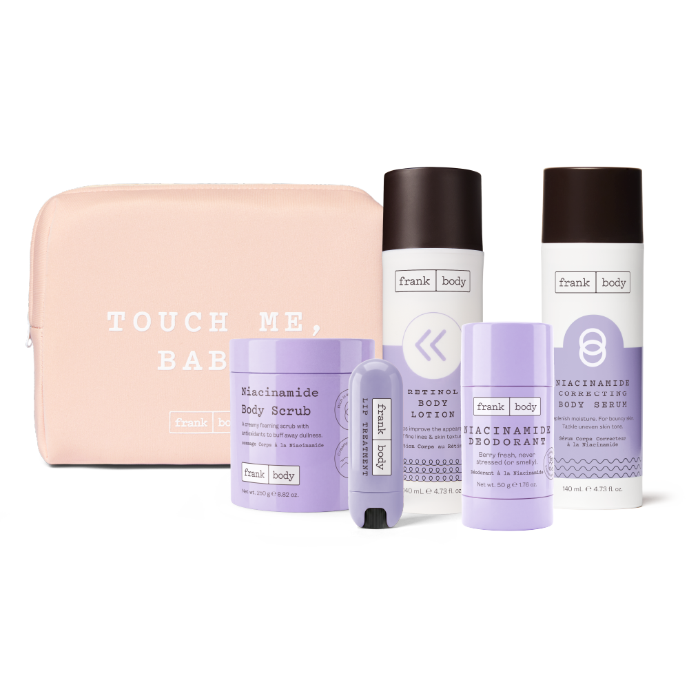 A Frank Body travel set featuring a light pink zippered pouch with white text, a purple body scrub jar, a white body lotion bottle, a deodorant stick, and a body serum bottle, all arranged on a white background.