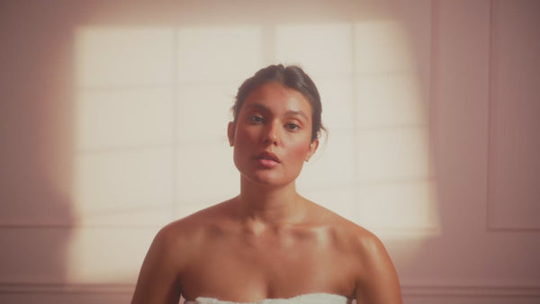 frank body Firm + Glow Tanning Jelly video: a woman wrapped in a towel in a sunlit room.